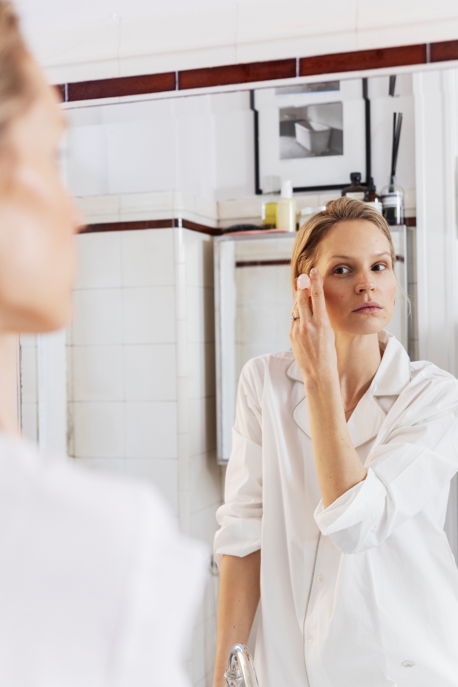 Woman doing her makeup in her bathroom, seen through the mirror and wearing white silk pyjamas.
